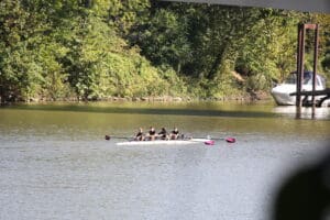 Four people rowing on a calm river, surrounded by lush greenery and a docked boat under a bridge.