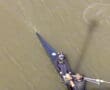 Aerial view of a three-person team rowing in a sleek boat on a river with brownish water, wearing black athletic gear.