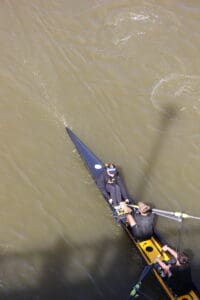 Aerial view of a three-person team rowing in a sleek boat on a river with brownish water, wearing black athletic gear.