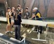 Rowing team receives coaching outdoors beside boats, preparing for a practice or competition.