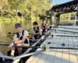 Rowing team on a sunny day at the dock, with a scenic bridge and trees in the background.