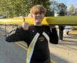 Rower gives thumbs up while holding a yellow boat on a sunny day. Dressed in NA Crew uniform.