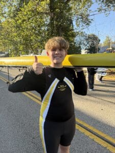 Rower gives thumbs up while holding a yellow boat on a sunny day. Dressed in NA Crew uniform.