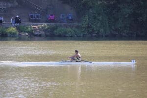 Single rower in a white boat on a calm river with spectators sitting by the shaded bank.