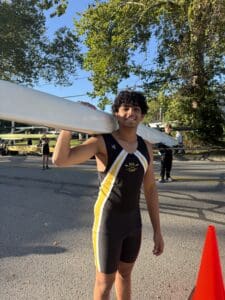 Rower carrying a boat on a sunny day, wearing a black and yellow team outfit, trees in the background.