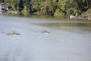 Rowing team gliding on a calm river surrounded by lush green trees and a riverside house.