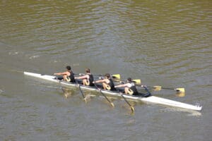 Four-person rowing team paddling in sync on a river, wearing black uniforms with yellow accents.
