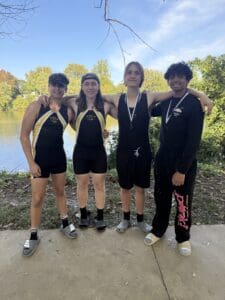 Four rowers in matching gear pose by a riverside with medals, surrounded by trees and a clear blue sky.