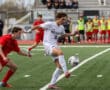 Soccer player in white jersey controls the ball during a match, with players in red and spectators in the background.
