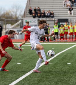 Soccer player in white jersey controls the ball during a match, with players in red and spectators in the background.