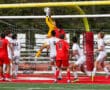 Soccer goalie leaps to catch the ball during a match, surrounded by players in red and white jerseys.