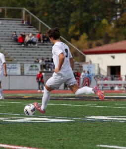 Soccer player in white kit dribbling the ball on a green field with spectators in the background.