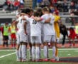 Soccer team huddle on field in white uniforms, preparing for a match in a stadium with spectators in background.