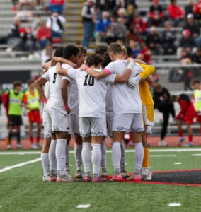 Soccer team huddle on field in white uniforms, preparing for a match in a stadium with spectators in background.