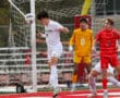 Soccer player heads the ball near goal, with teammates and goalkeeper watching, during an intense match.