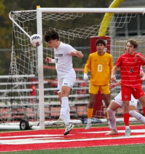 Soccer player heads the ball near goal, with teammates and goalkeeper watching, during an intense match.