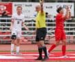 Soccer referee issues yellow card during high school match, players react on field.