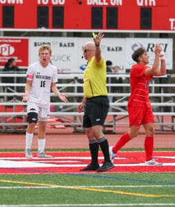 Soccer referee issues yellow card during high school match, players react on field.