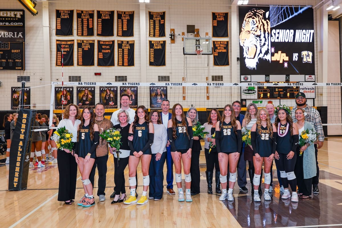 Volleyball team celebrating senior night with coaches and family on the court. Banners and scoreboard in the background.