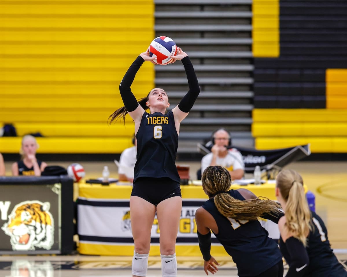 Volleyball player setting ball during a match in gym, wearing black jersey with number 6.