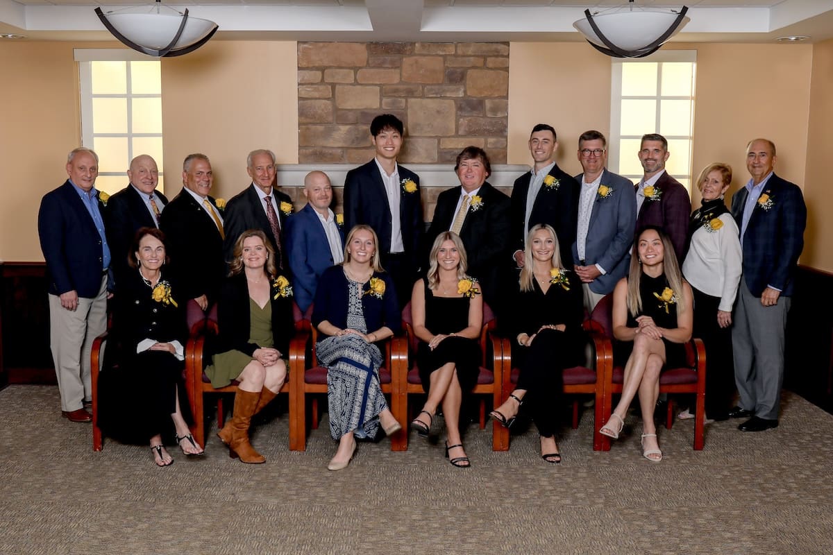 Group photo of 18 people dressed formally, wearing yellow corsages, sitting and standing in a room with a stone wall background.