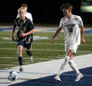 Two soccer players in action on a field at night, one in a white uniform, the other in a blue and gold uniform.