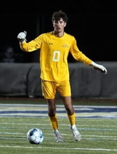 Soccer goalie in yellow kit prepares for a goal kick during a night match on a field.