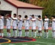 Soccer team in white kit stands in unity during pre-game ceremony on field with red-roofed building in background.