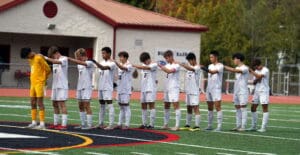 Soccer team in white kit stands in unity during pre-game ceremony on field with red-roofed building in background.
