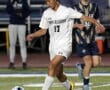 Soccer player in white uniform dribbles ball during a high school match, focused and in motion on the field.