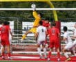 Soccer goalie in yellow jumps to catch ball during intense match with players in red and white jerseys.