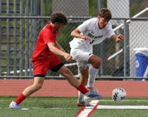 Two soccer players competing for the ball on a field during a match, one in red and the other in white.
