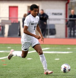 Soccer player in white jersey dribbles on the field during a match, showcasing action and focus.