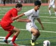 Two soccer players compete for the ball during a match on a green field. One is wearing white, and the other is in red.