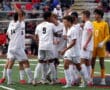 High school soccer team huddles on field in white jerseys, preparing for game. Goalkeeper in yellow.
