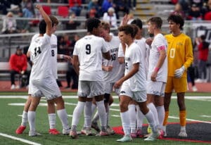 High school soccer team huddles on field in white jerseys, preparing for game. Goalkeeper in yellow.