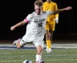 Soccer player in white jersey kicks ball mid-action during nighttime match, with player in yellow in background.