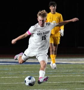 Soccer player in white jersey kicks ball mid-action during nighttime match, with player in yellow in background.