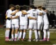 Soccer team huddles together on the field in white uniforms during a nighttime match.