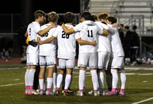 Soccer team huddles together on the field in white uniforms during a nighttime match.