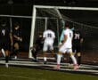 Soccer players in black and white jerseys around the goalpost during a nighttime match.