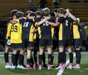 Soccer team in a huddle on the field, wearing black and yellow uniforms with pink socks, ready for the match.