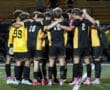 Soccer team huddling together on the field, wearing black and yellow uniforms, preparing for the match.