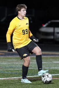 Soccer goalie in yellow jersey and gloves controls the ball during a night game on turf field.