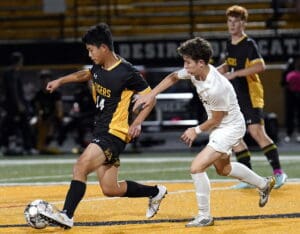 Soccer players competing in a match, one in black-gold uniform, the other in white.