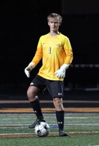 Young soccer goalkeeper in yellow jersey prepares to kick ball on sports field at night.