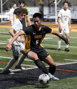 Soccer player in black and yellow jersey dribbles ball past opponent on a sports field at night.