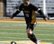 Soccer player in black and gold uniform dribbles ball on field during a night game.