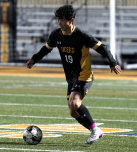 Soccer player in black and gold uniform dribbles ball on field during a night game.