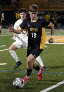 Soccer player in black and yellow uniform dribbles past opponent on a field during a night match.